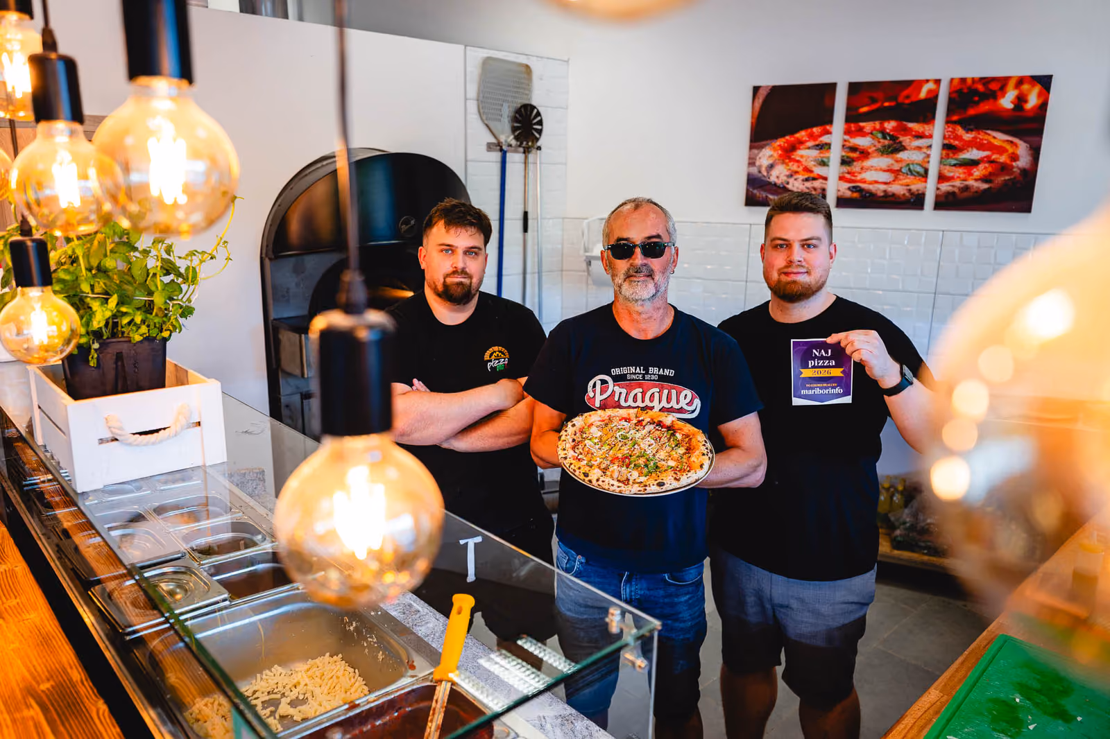 Miran Gomboc mit seinen beiden Söhnen hinter dem Tresen in Lenart, in der Hand eine frisch gebackene Pizza und die Auszeichnung „Naj pizza 2026“